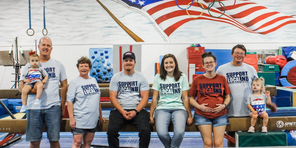 A group photo of the family that owns J and R Gymnastics, taken in a gymnastics facility. Seven people of various ages are seated on a gymnastics beam, all wearing "Support Local NB" T-shirts. Behind them, colorful gymnastics equipment and an American flag mural are visible.